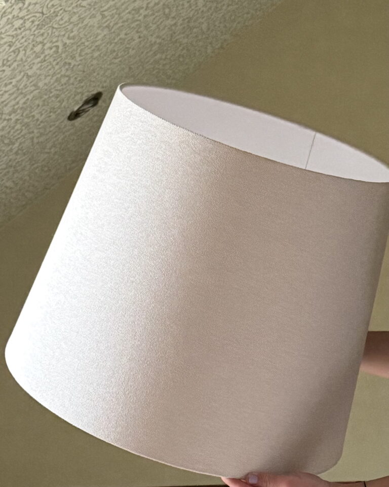 Hand holding beige textured linen lampshade, angled upward against a soft wall background